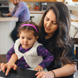 woman with a child on her lap, at her kitchen counter on a laptop