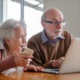Older adults sitting at a table, looking at a laptop while one person holds a smartphone in a home setting.