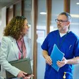 Two people with ID badges walk through a medical office hallway while talking.