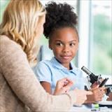 Instructor guides a girl during a classroom science activity using a microscope.