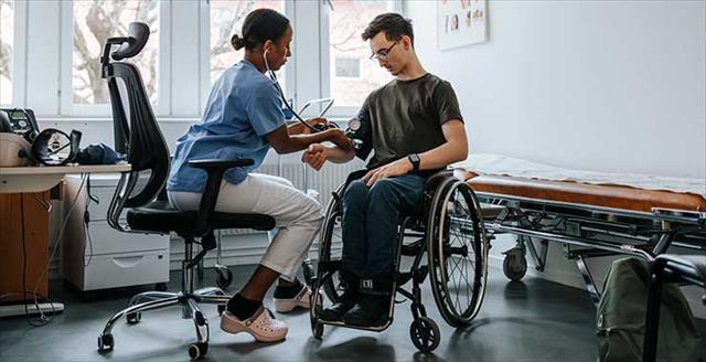 Doctor checking blood pressure of young male sitting on wheelchair at examination room at clinic