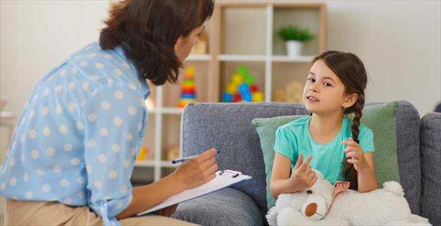 Happy little girl telling story to child psychologist during therapy session in cozy modern office