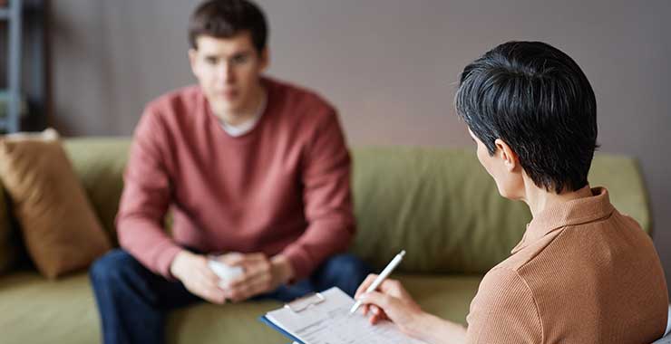 Young man sitting on couch as therapist takes notes