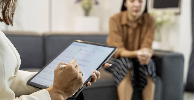 Clinician holds a digital tablet while a patient sits on a sofa.