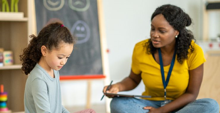 A mental health specialist holding a clipboard sits beside a young girl in a classroom, observing her