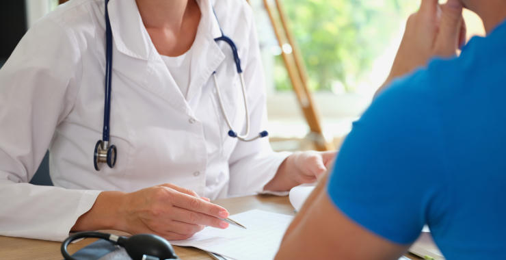 Healthcare professional with a stethoscope consulting with a patient across a desk in a medical office.