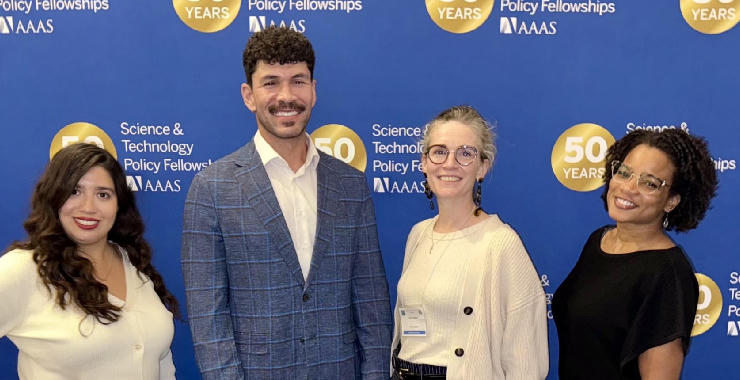 Rubi Gonzales, Steven W. Kasparek, Molly Below, and Natalie Anumba standing together in front of a banner celebrating the 50th anniversary of the Science & Technology Policy Fellowships.