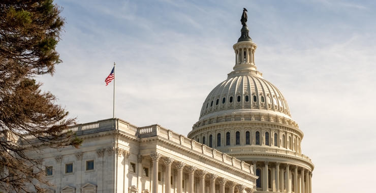 View of the U.S. Capitol building dome and American flag against a clear sky.