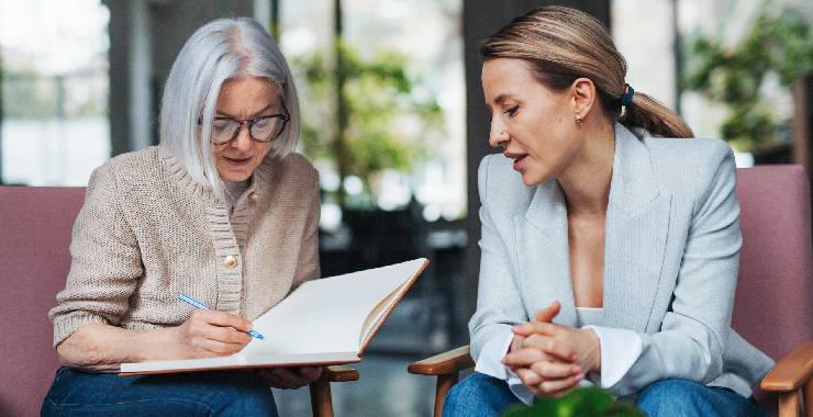 Two women sit side by side closely reviewing notes in an open notebook.
