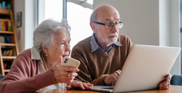 Older adults sitting at a table, looking at a laptop while one person holds a smartphone in a home setting.