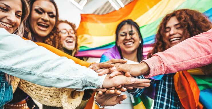 Diverse group of people standing together with hands stacked in the center beneath a rainbow pride flag.