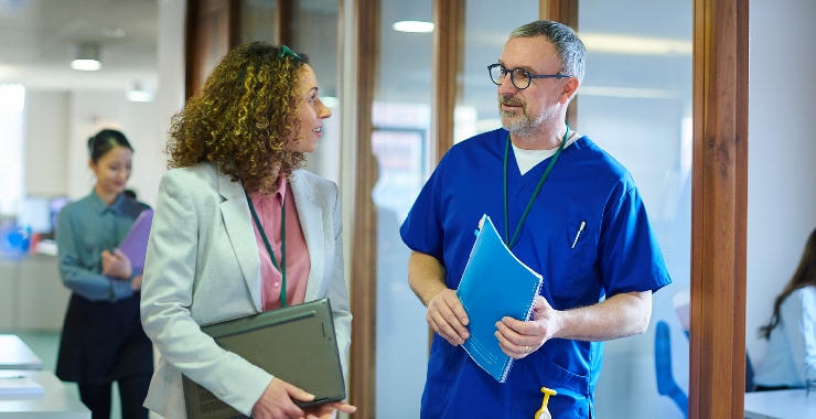 Two people with ID badges walk through a medical office hallway while talking.