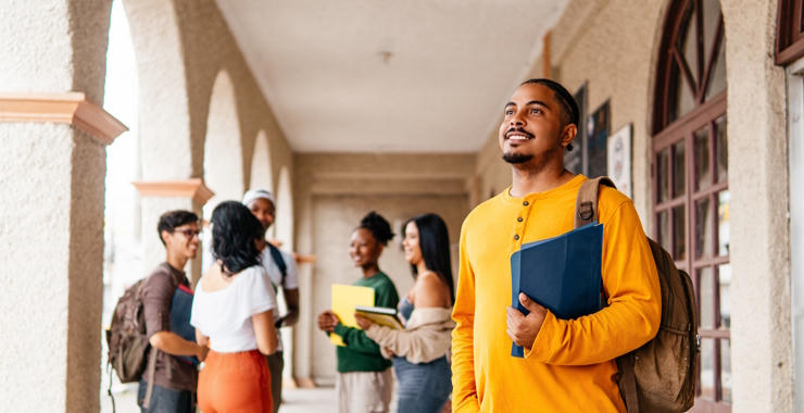 Students gather and talk in a campus walkway while a student in the foreground carries folders and a backpack.
