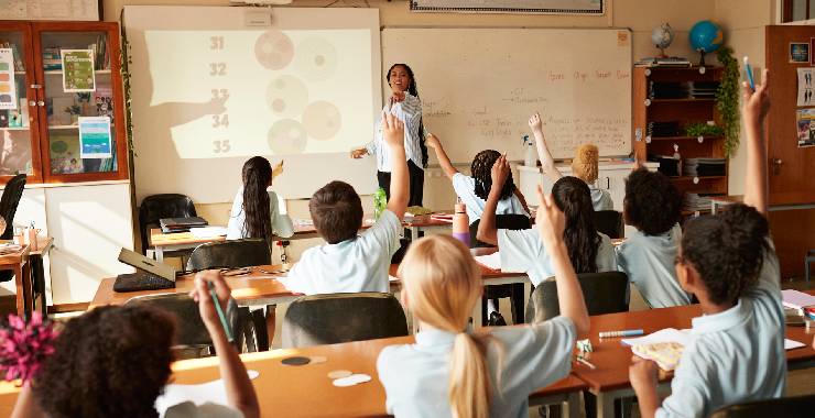 Teacher stands at front of classroom with students raising hands to answer a question.