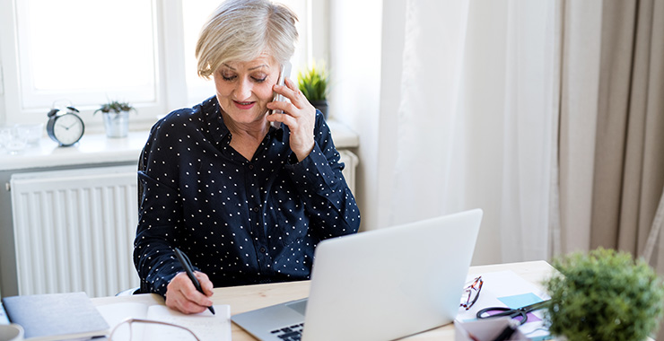 Lady multitasking on cell phone, writing in front of laptop.